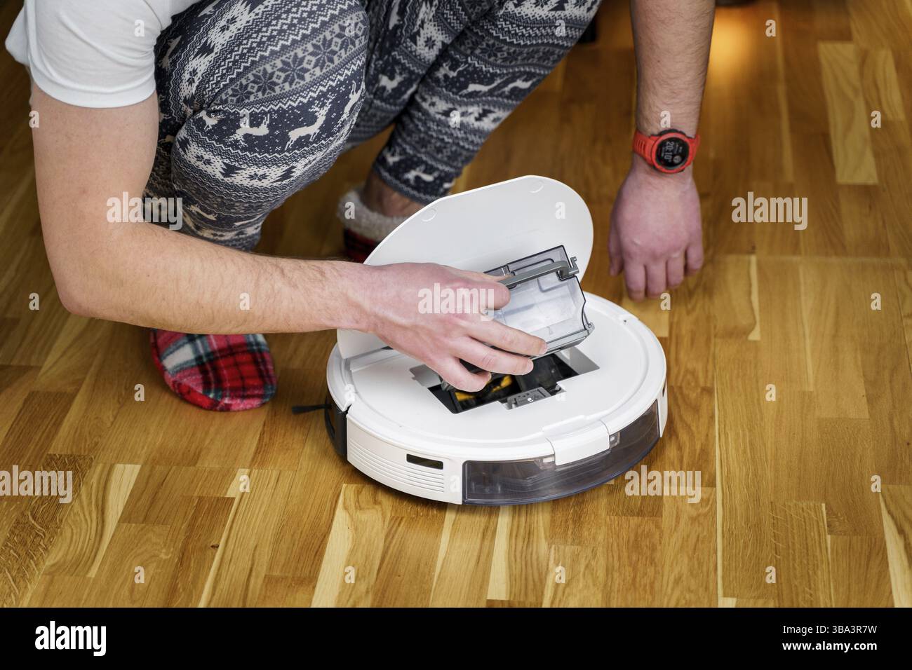 Kaukasischen jungen Mann in weihnachten Pyjamas tun Staubsauger Roboter Wartung. Männlich im Winter gemusterte Nachtwäsche und rote Socken Hausschuhe beginnt automatisch Stockfoto
