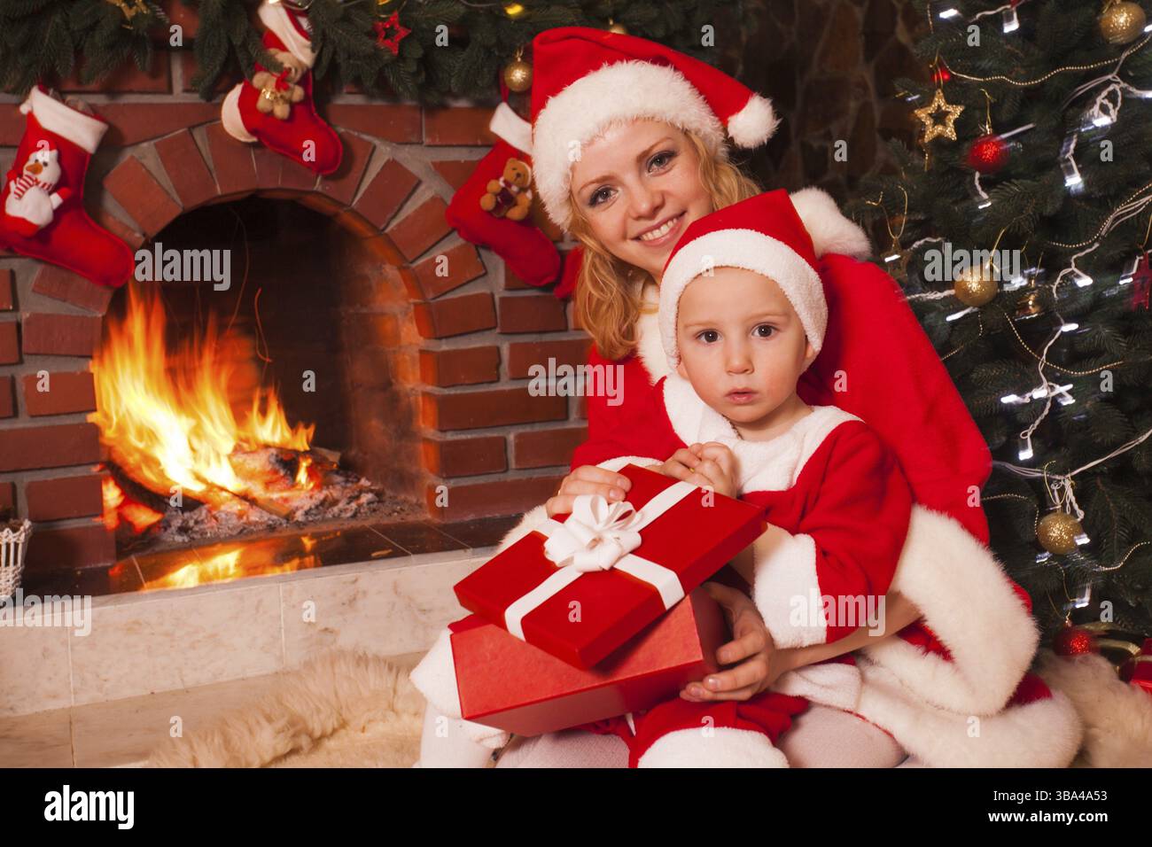 Mutter und Sohn sitzen neben Kamin und weihnachtsbaum. Die Familie schaut in eine Geschenkbox Stockfoto
