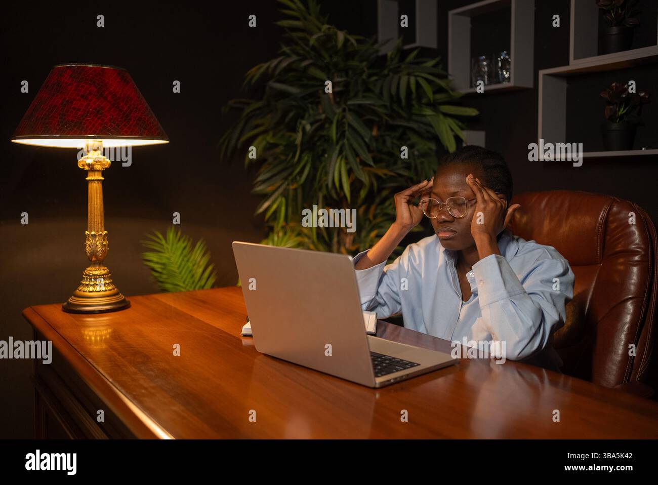 Professionelle afroamerikanische Arbeiterin, die unter intensivem Stress leidet und die Fingerspitzen gegen die Stirn drückt, während sie bis spät unter weichem Lampenlicht arbeitet, und der Laptop-Bildschirm wirft Schatten Stockfoto