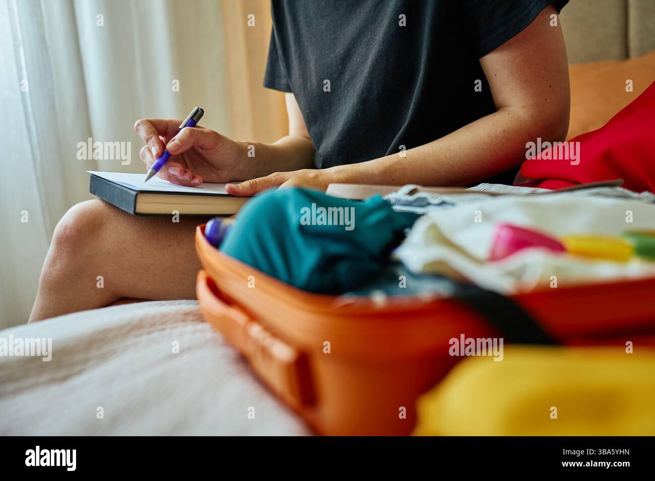 Die Person schreibt in einem Notizbuch mit einem orangen Koffer voller Kleidung. Reisekonzept mit handschriftlicher Checkliste Stockfoto