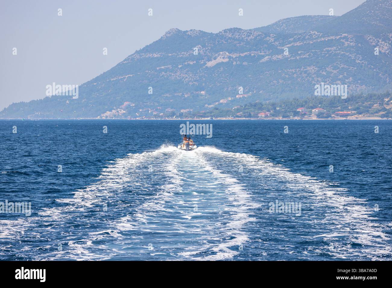 Ein Boot fährt über das Meer und hinterlässt eine weiße Spur. Zwei Personen sind an Bord. In der Ferne erheben sich Berge über dem Wasser, und eine kleine Stadt ist zu sehen Stockfoto