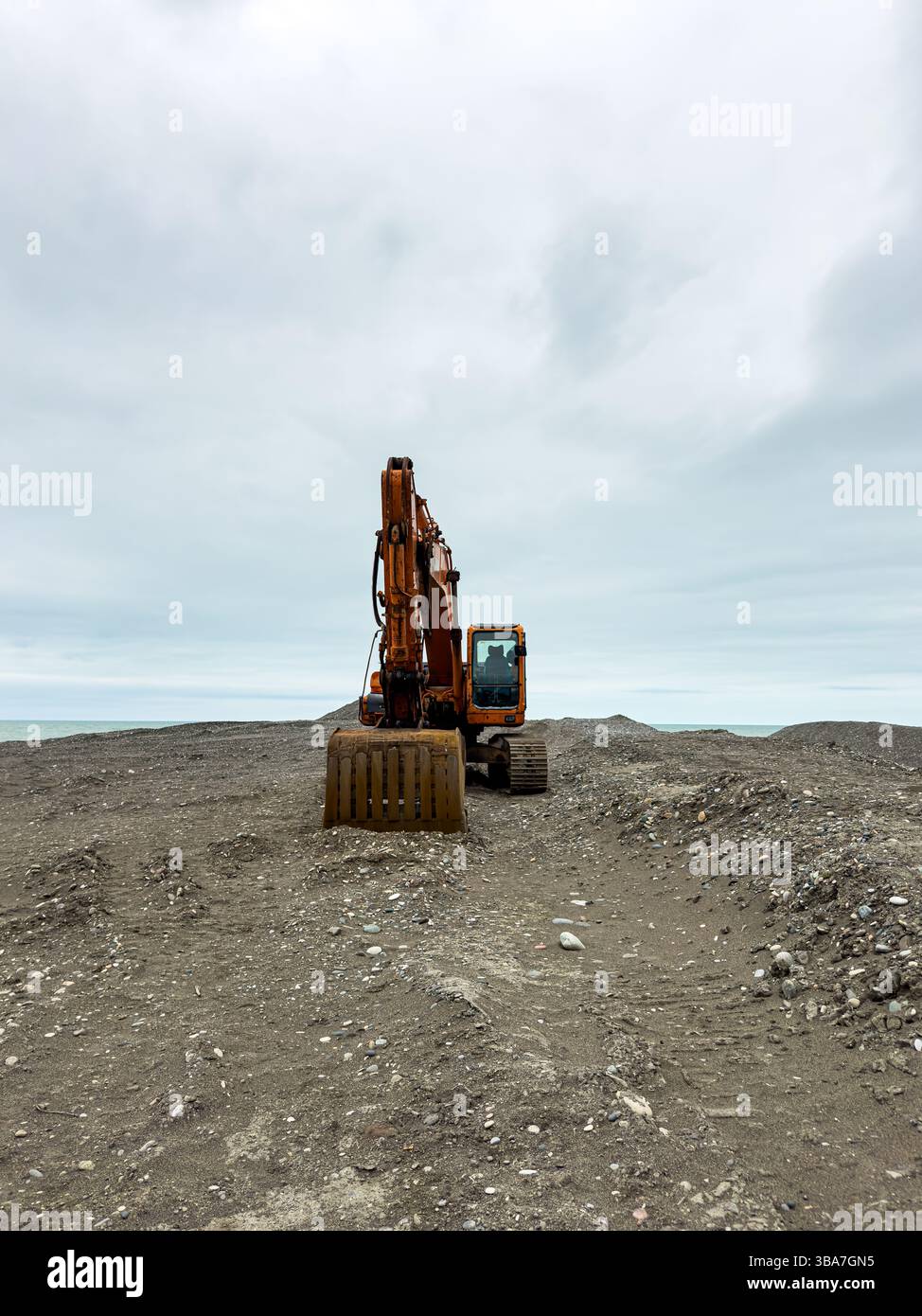 Orangefarbener Bagger auf leerem Küstengelände unter bedecktem Himmel. Zentrierter Blick, bewölktes Wetter, keine Leute. Industrie, Erdbewegung, Baukonzept. Stockfoto