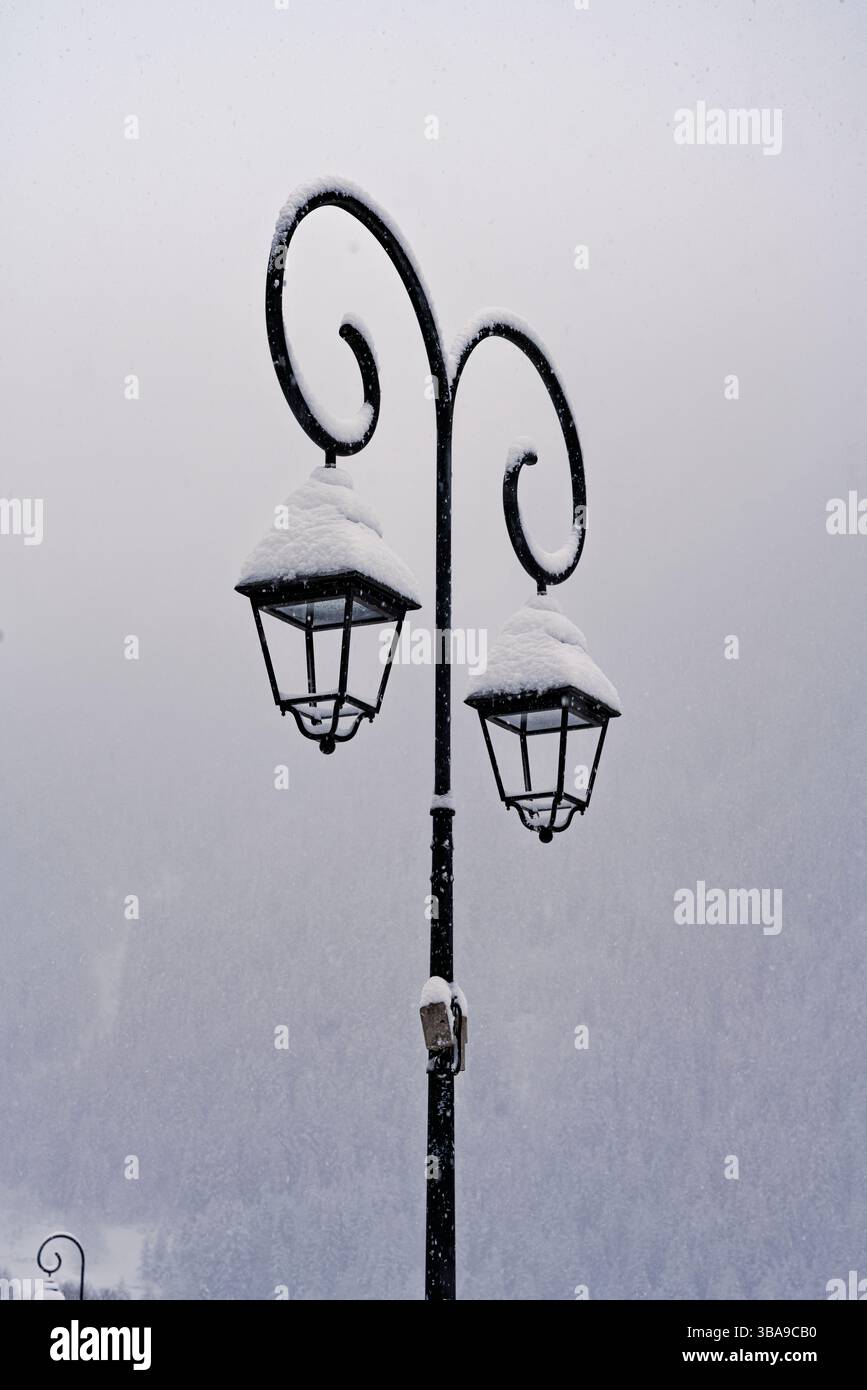 Eine doppelte Straßenlaterne krümmt sich anmutig in den weißen Himmel, der schwarze Eisenrahmen wird von perfekten Schneekuppeln in Peisey bedeckt. Stockfoto