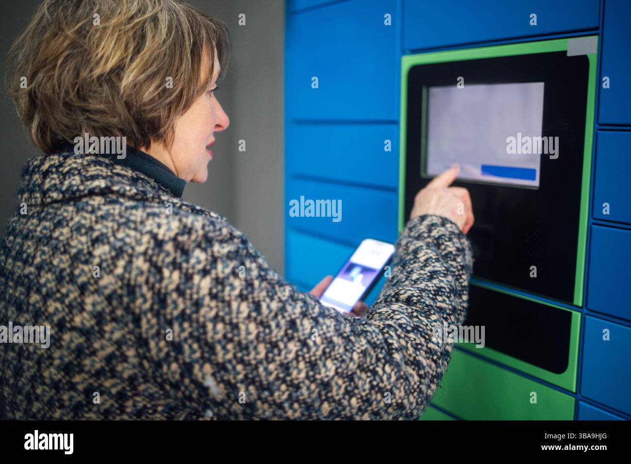 Eine ältere Frau tippt auf das Display, um ein Paket zu erhalten. Eine reife Dame in lässiger Kleidung gibt Daten auf einem Paketautomaten ein. Der Rentner hält das Telefon Stockfoto