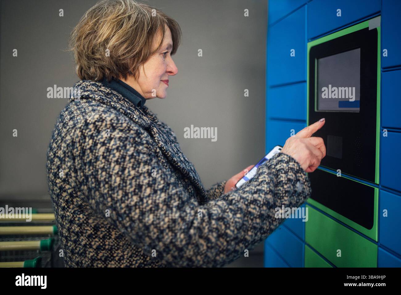 Eine ältere Frau tippt auf das Display, um ein Paket zu erhalten. Eine reife Dame in lässiger Kleidung gibt Daten auf einem Paketautomaten ein. Der Rentner hält das Telefon Stockfoto