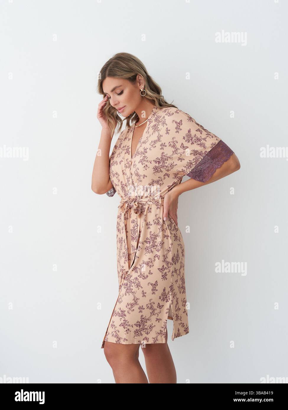 Frau in einem hellen Blumenkleid mit Bindegürtel und lila Ärmeln posiert nachdenklich und berührt ihr Haar. Sie wirkt ruhig und introspektiv Stockfoto