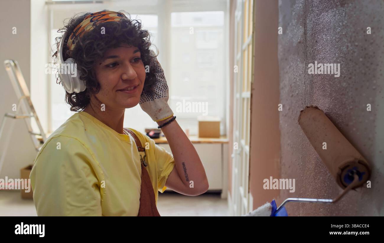 Hispanische Frau mit Kopfhörern und Handschuhen, die eine Innenwand mit einer Rolle bemalt. Hintergrund zeigt Werkzeuge und natürliches Licht, das durch Fenster strömt Stockfoto Hispanische Frau mit Kopfhörern und Handschuhen, die eine Innenwand mit einer Rolle bemalt. Hintergrund zeigt Werkzeuge und natürliches Licht, das durch Fenster strömt Stockfoto