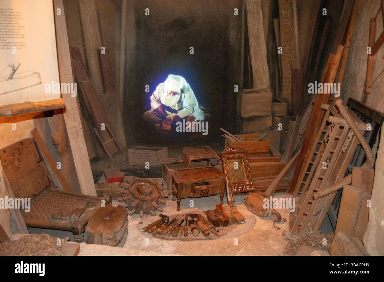 Ein schwach beleuchteter Raum mit verschiedenen antiken Werkzeugen und Holzkisten. Im Hintergrund wird eine geisterhafte Figur projiziert, die lesend oder schreibend wirkt. Die Stockfoto