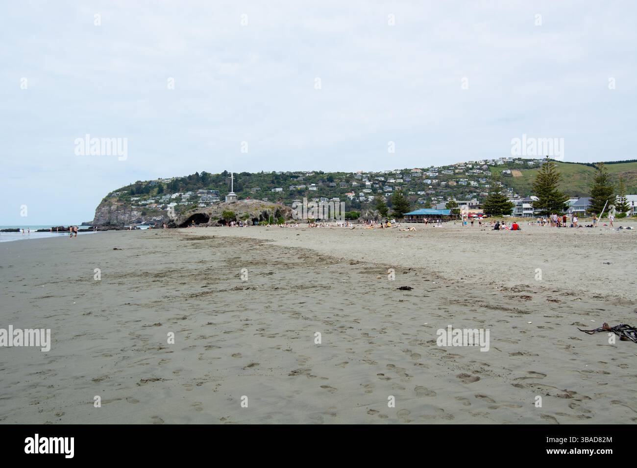 Ein Sandstrand mit Blick auf einen Hügel im Hintergrund, mit Häusern gesäumt. Die Menschen sind am Strand verstreut und genießen den Tag. Der Himmel ist bedeckt Stockfoto