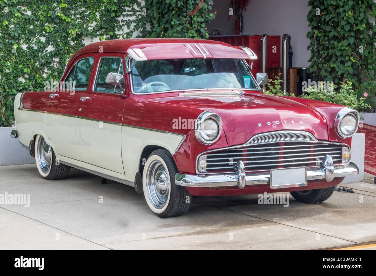 Ein klassischer zweifarbiger Ford Zodiac-Wagen in Rot und Creme parkt auf einer Betoneinfahrt neben einem mit grünen Kletterpflanzen bedeckten Gebäude. Der Vintage Stockfoto