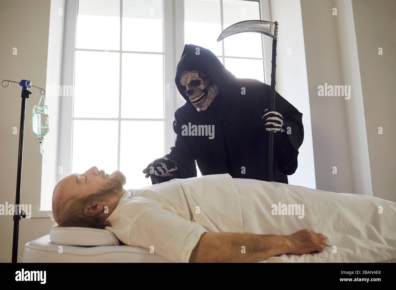 Die todhaltige Sense steht neben dem sterbenden älteren Mann, der in der medizinischen Klinik unter IV-Tropf liegt. Stockfoto