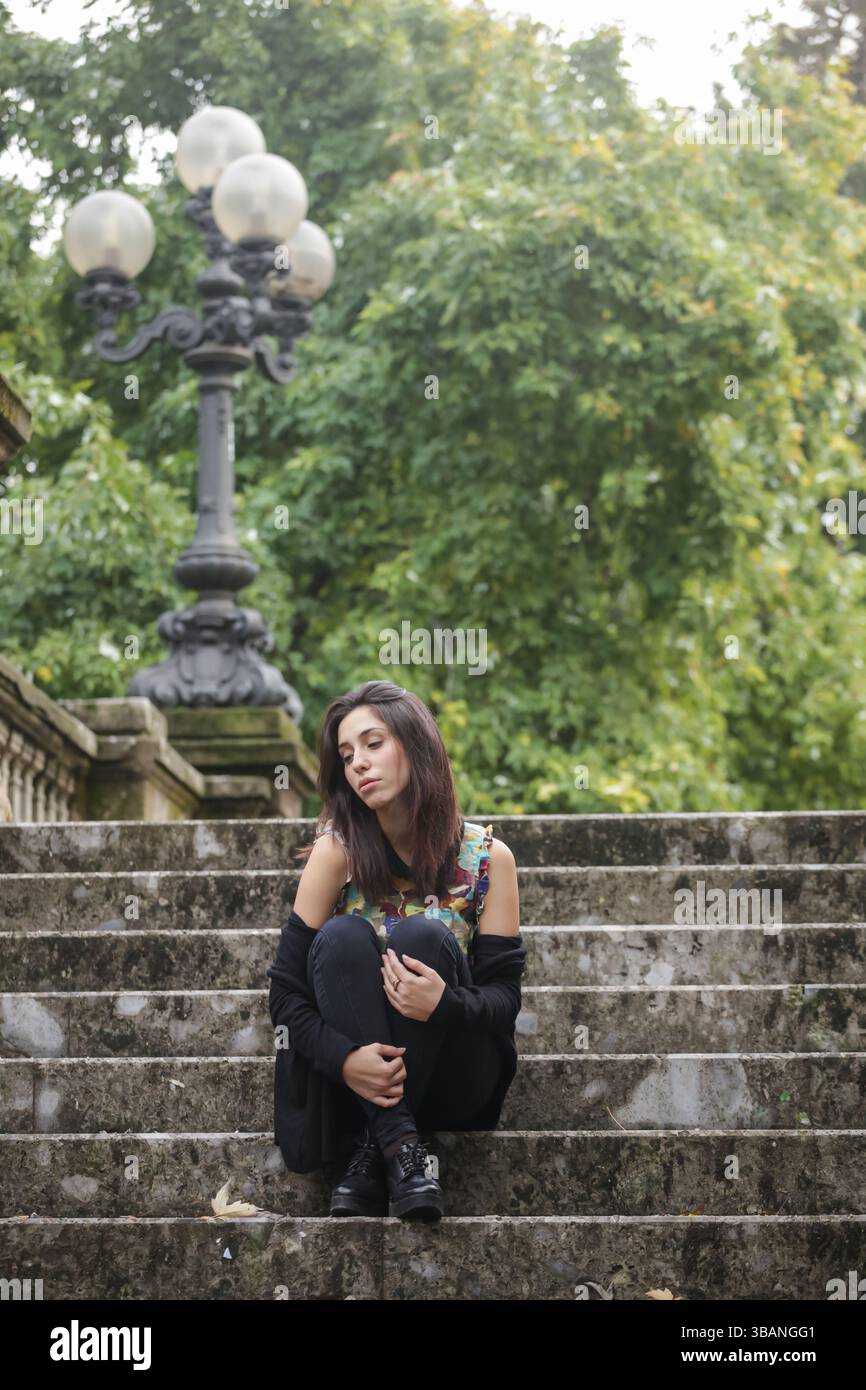 Junge Frau, die in nachdenklicher Stimmung auf Steintreppen sitzt, ein farbenfrohes ärmelloses Oberteil und eine schwarze Jeans trägt. Stockfoto
