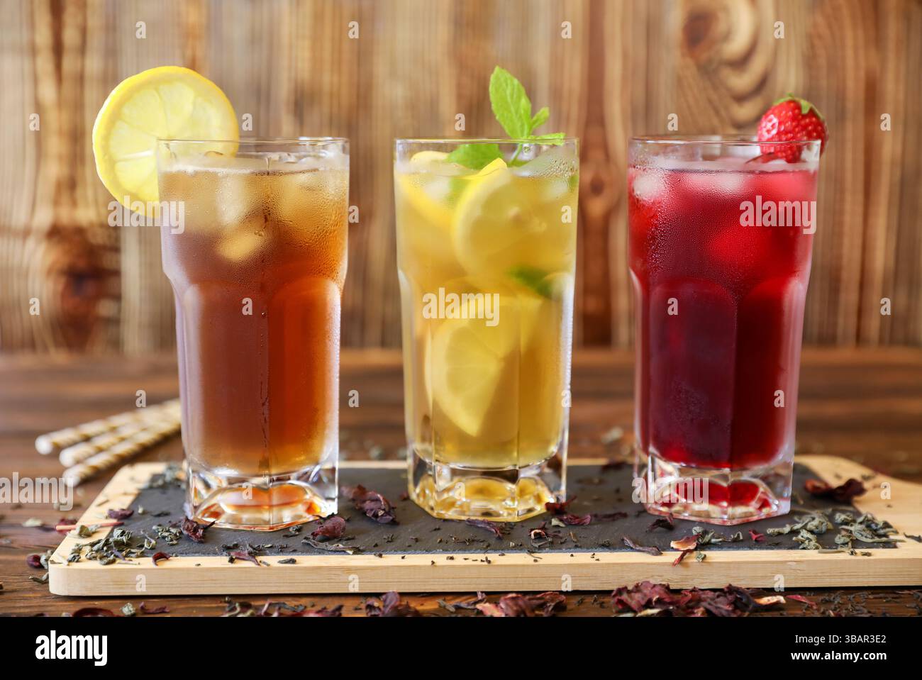 Auf einem rustikalen Holztisch stehen drei große Gläser mit Eistee, Limonade mit Zitronenscheiben und ein Beerengetränk mit Kirschen. Frische Minzgarnituren sorgen für einen lebendigen Touch. Stockfoto