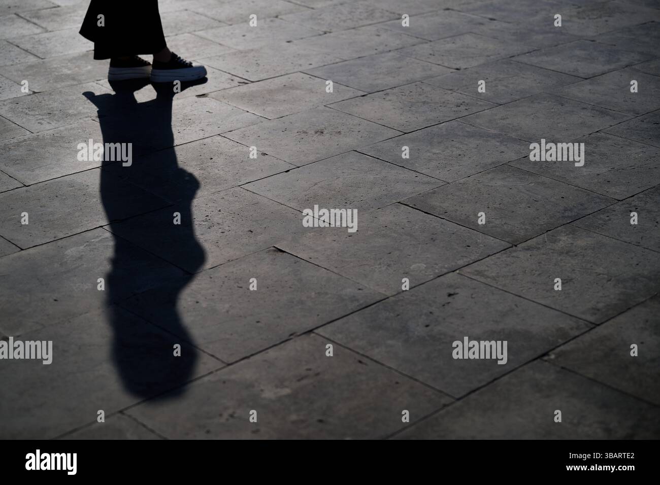 Die partielle Silhouette einer Person, die auf einem Bürgersteig läuft, erzeugt ein surreales Spiel zwischen Körper und Schatten und bildet abstrakte Formen. Stockfoto