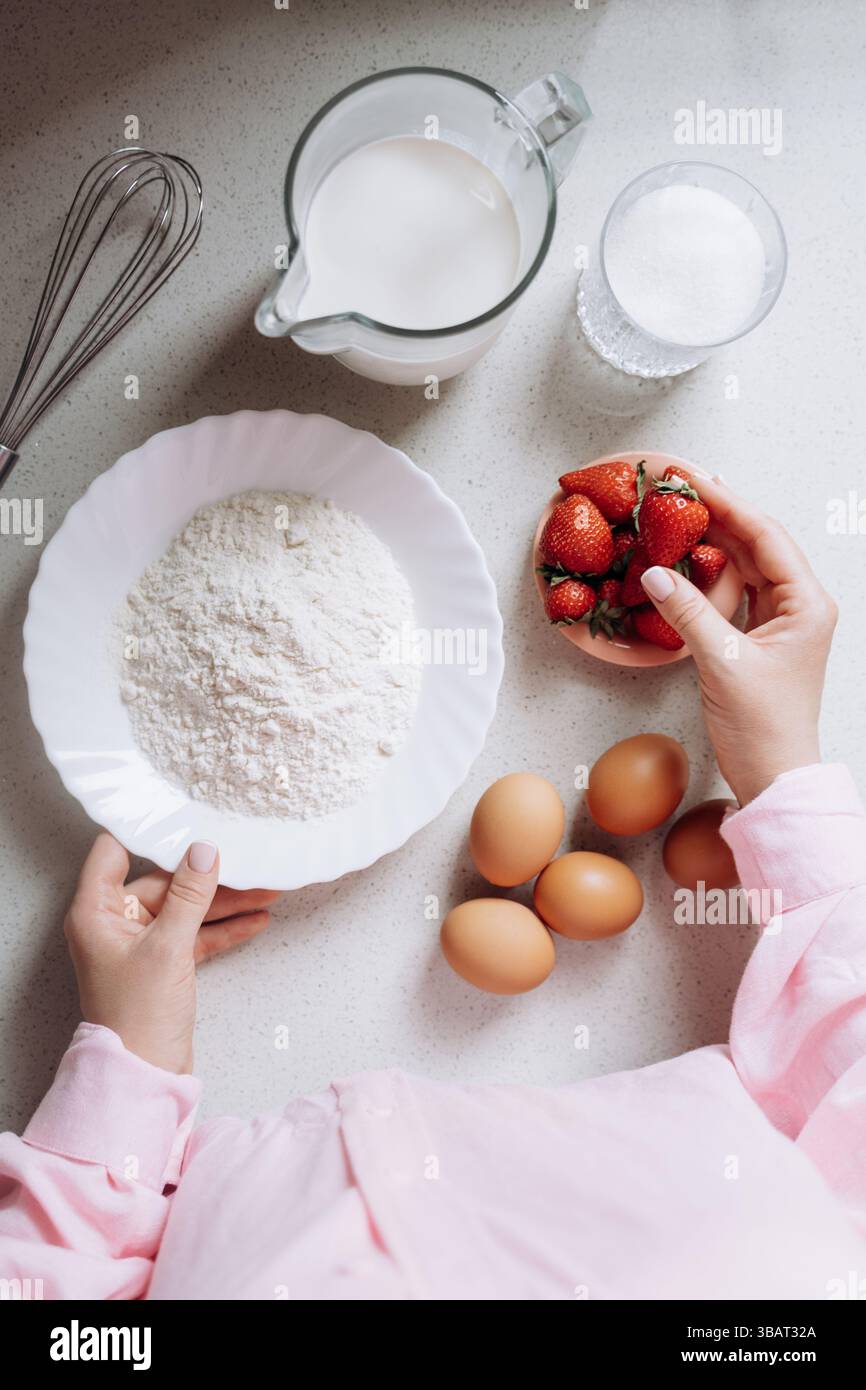 Die Zutaten zum Backen sind auf einer Küchentheke ordentlich angeordnet, einschließlich Mehl, Zucker, Milch, Eier, und frische Erdbeeren. Die Hände sind abgebildet, die einen halten Stockfoto