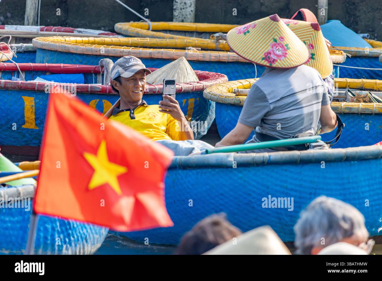 HOI AN, VIETNAM, 26. April 2025, der geschickte Steuermann eines traditionellen Rundbootes lehnt sich über das Wasser und macht Fotos von Touristen Stockfoto