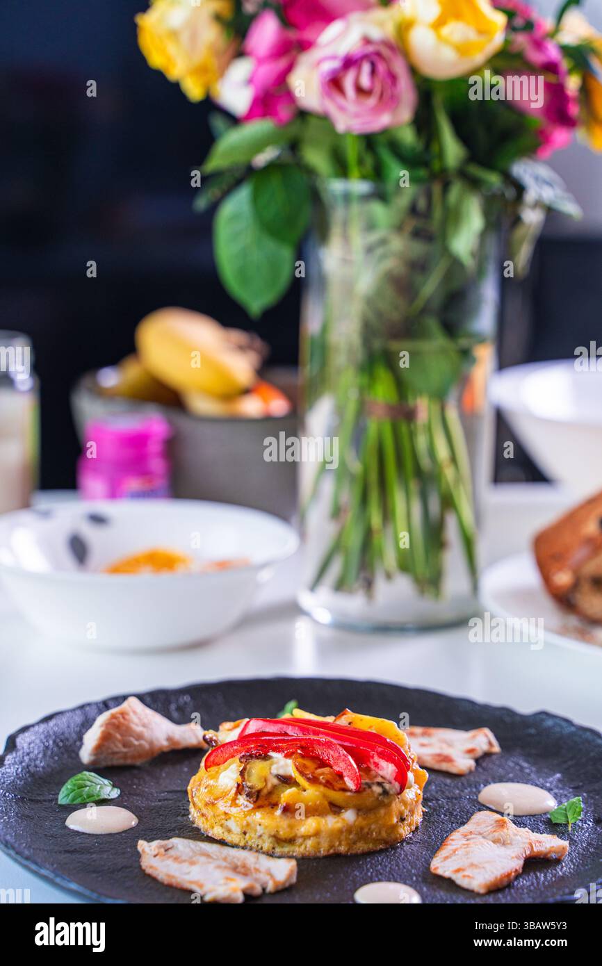 Romantisches Galadinner. Gekochte Hühnchen- oder putenstücke mit Kartoffeltartilla und rotem Paprika auf einem schwarzen Teller auf dem Tisch mit einem Strauß Rosen. Hg Stockfoto