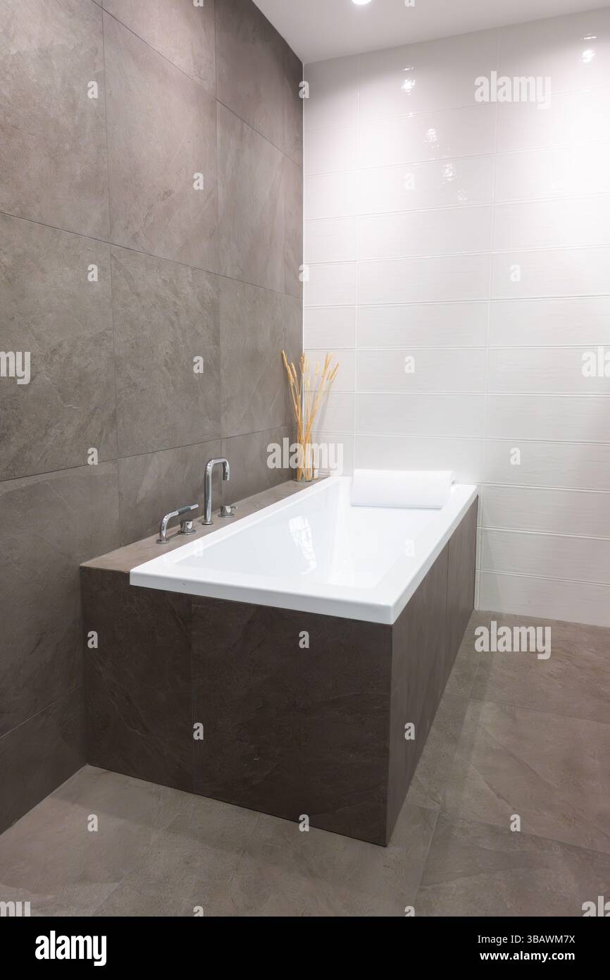 Moderne eingebaute Keramikbadewanne mit Edelstahl- und Chromarmaturen in einem modernen Badezimmer mit grauen Fliesenwänden und Boden für ein elegantes Zuhause Stockfoto