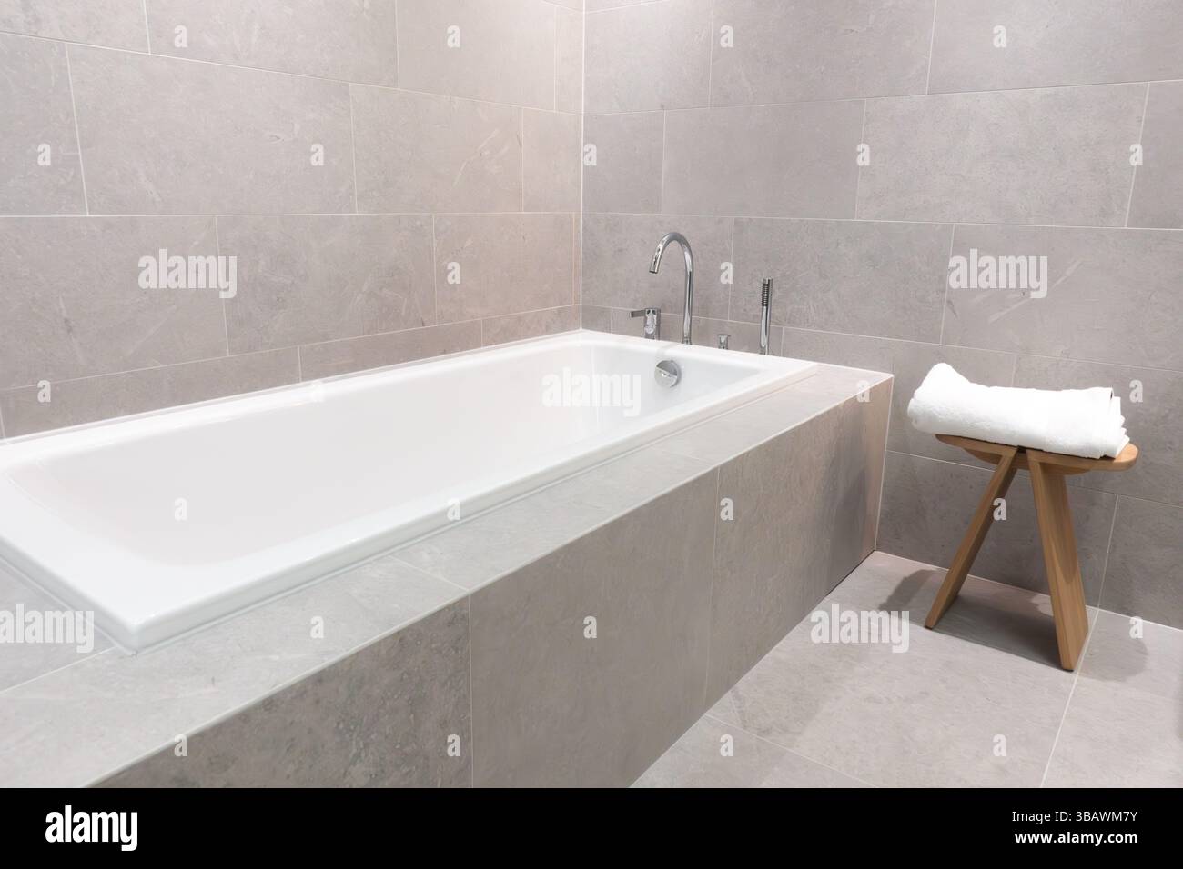 Moderne eingebaute Keramikbadewanne mit Edelstahl- und Chromarmaturen in einem modernen Badezimmer mit grauen Fliesenwänden und Boden für ein elegantes Zuhause Stockfoto