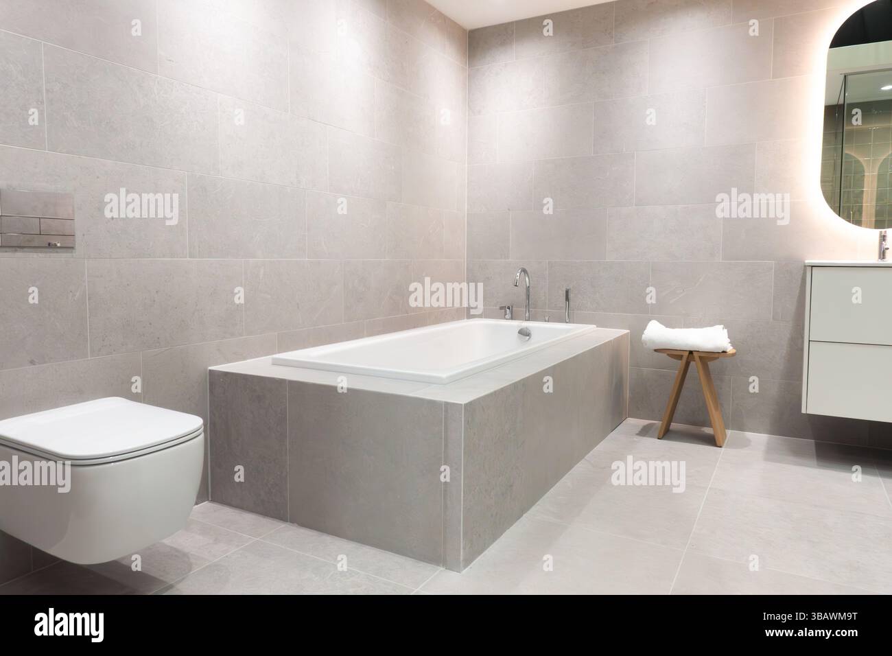 Moderne eingebaute Keramikbadewanne mit Edelstahl- und Chromarmaturen in einem modernen Badezimmer mit grauen Fliesenwänden und Boden für ein elegantes Zuhause Stockfoto
