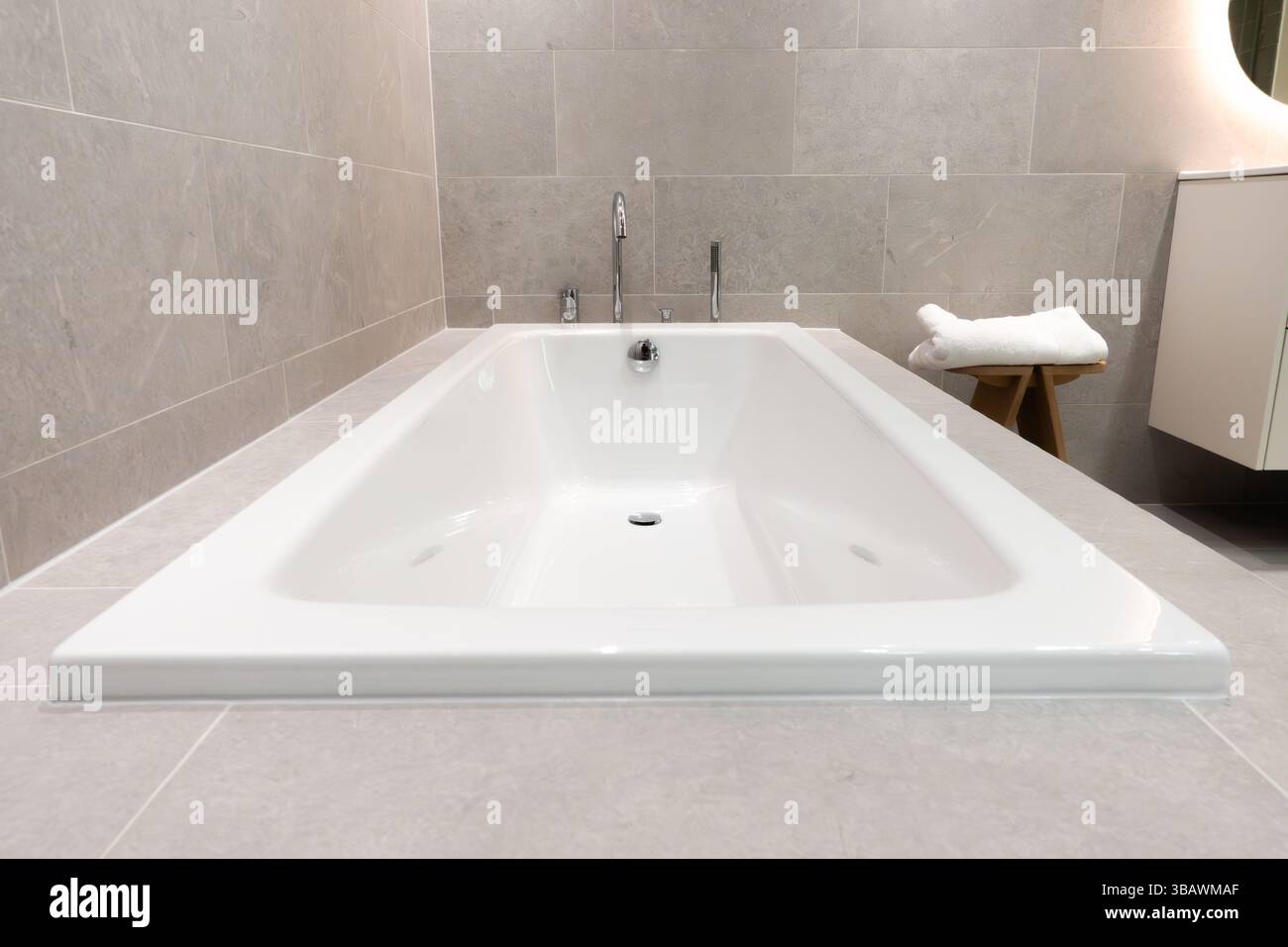 Moderne eingebaute Keramikbadewanne mit Edelstahl- und Chromarmaturen in einem modernen Badezimmer mit grauen Fliesenwänden und Boden für ein elegantes Zuhause Stockfoto