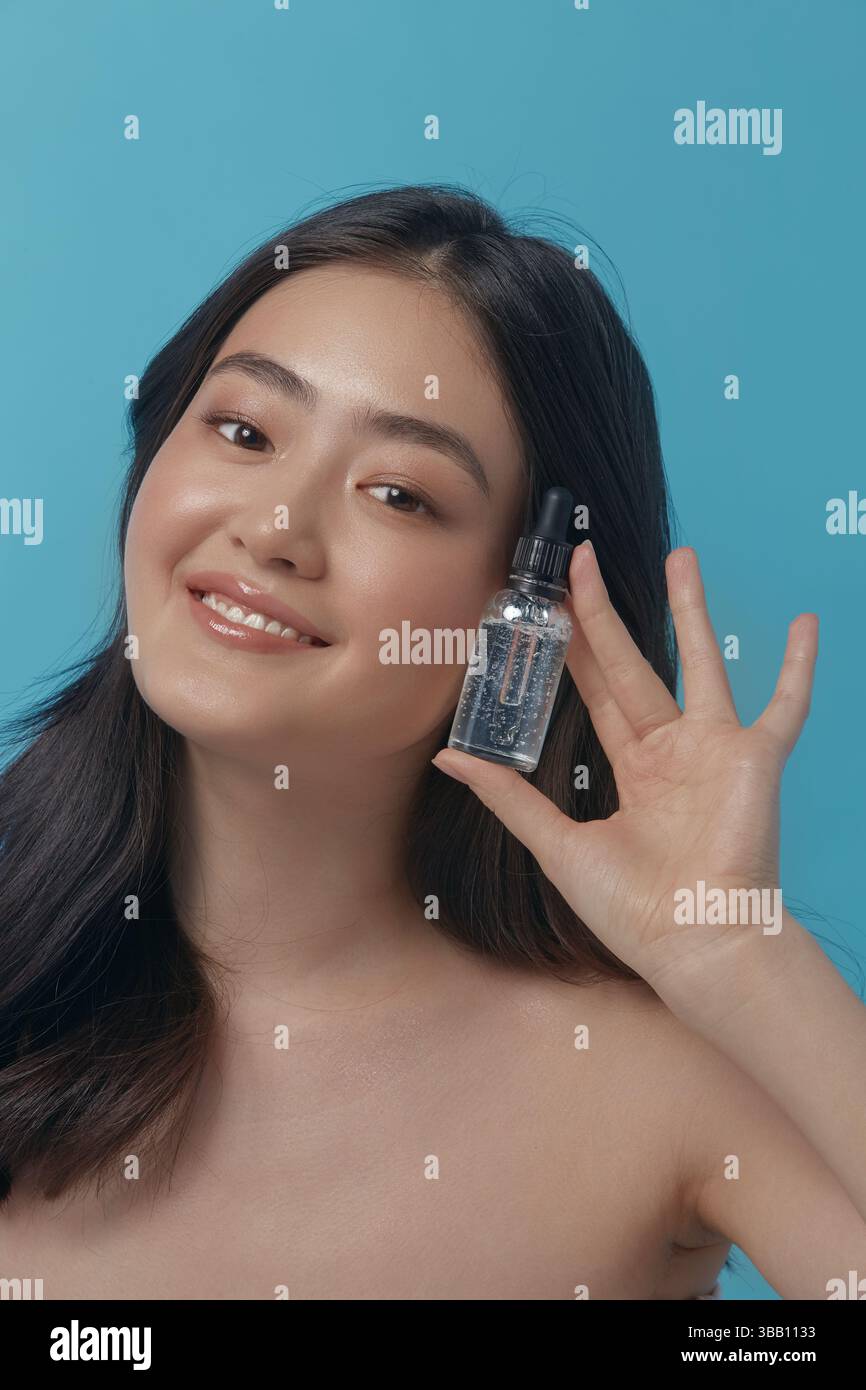 Fröhliche japanische Ethnicity-Frau, die ein Gesichtsserum in der Nähe des Selbstgesichts hält, drückt Freude und Freude aus, posiert auf blauem Hintergrund, Nahaufnahme senkrecht Stockfoto