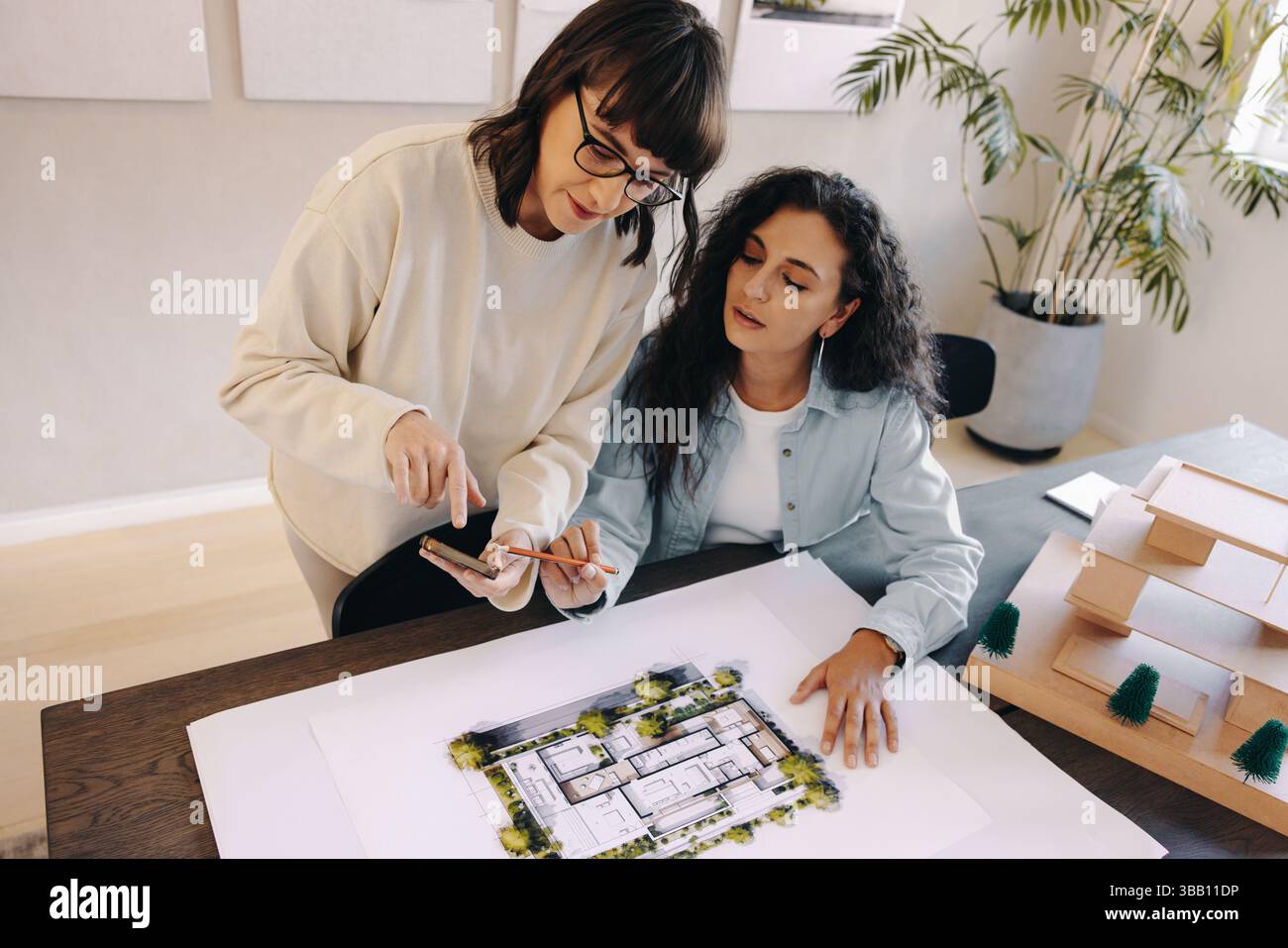 Zwei Frauen arbeiten in einem Architekturbüro zusammen, eine zeigt Pläne und nutzt ein Smartphone für Diskussionen, um Teamarbeit zu fördern. Stockfoto