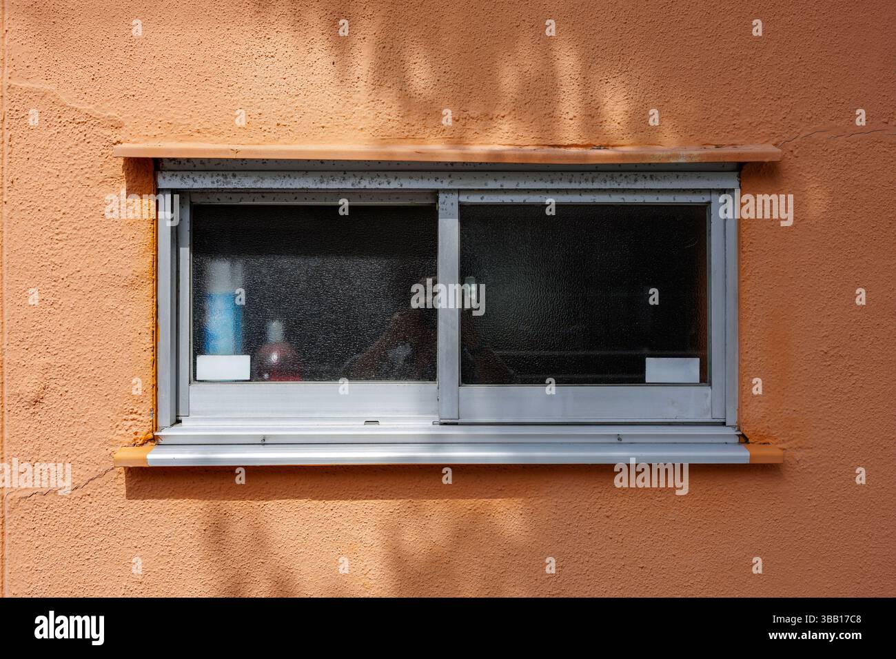 Rechteckiges Glasfenster in orangefarbener Stuckwand. Umliegende Bäume erzeugen einen Yin-Yang-Schatten, der Zen-ähnliche Ruhe schafft. Stockfoto