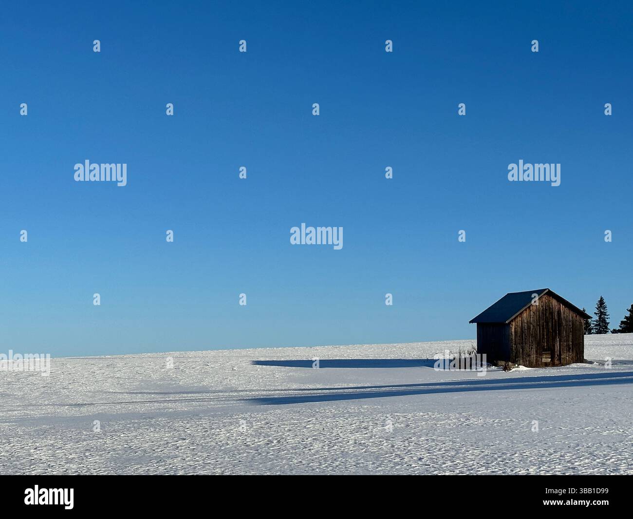 Eine rustikale Holzscheune steht allein auf einem schneebedeckten Feld unter einem riesigen, klaren blauen Himmel. - Smartphone-aufgenommenes Stockfoto