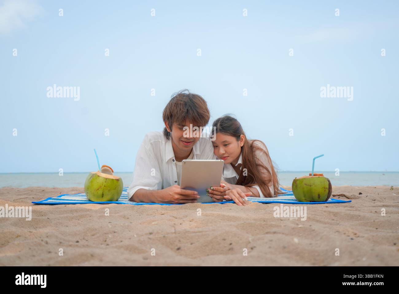 Junges asiatisches Paar, das am Strand liegt und einen entspannenden Moment genießt und Inhalte auf einem digitalen Tablet ansieht, mit Kokosnussgetränken auf jeder Seite. Ein gemütliches und Stockfoto