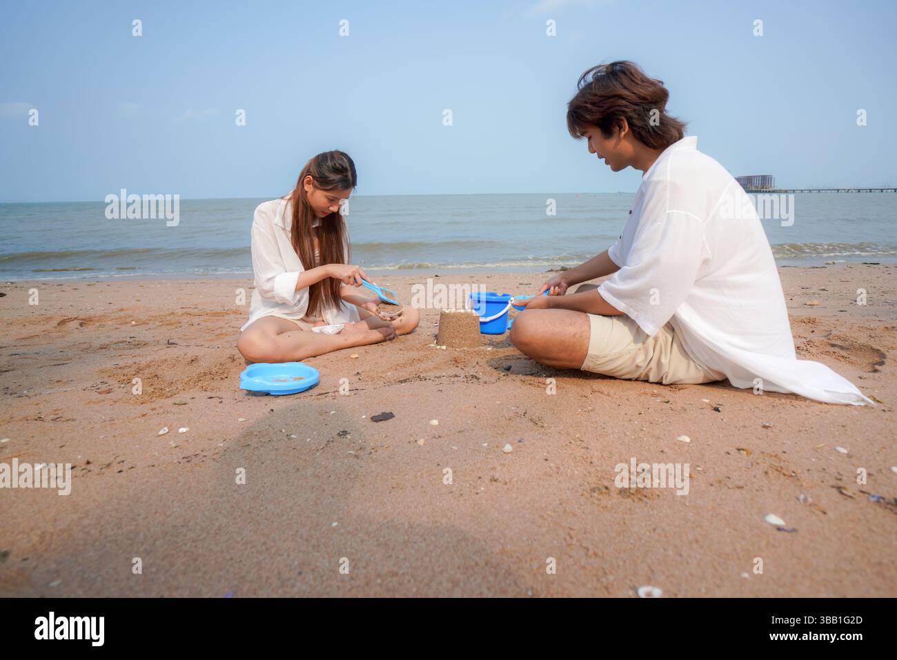 Junge asiatische Paare genießen spielerische Zeit am Strand, während sie zusammen eine Sandburg bauen, umgeben von tropischer Landschaft und ruhigem Meer, die einen SENS weckt Stockfoto