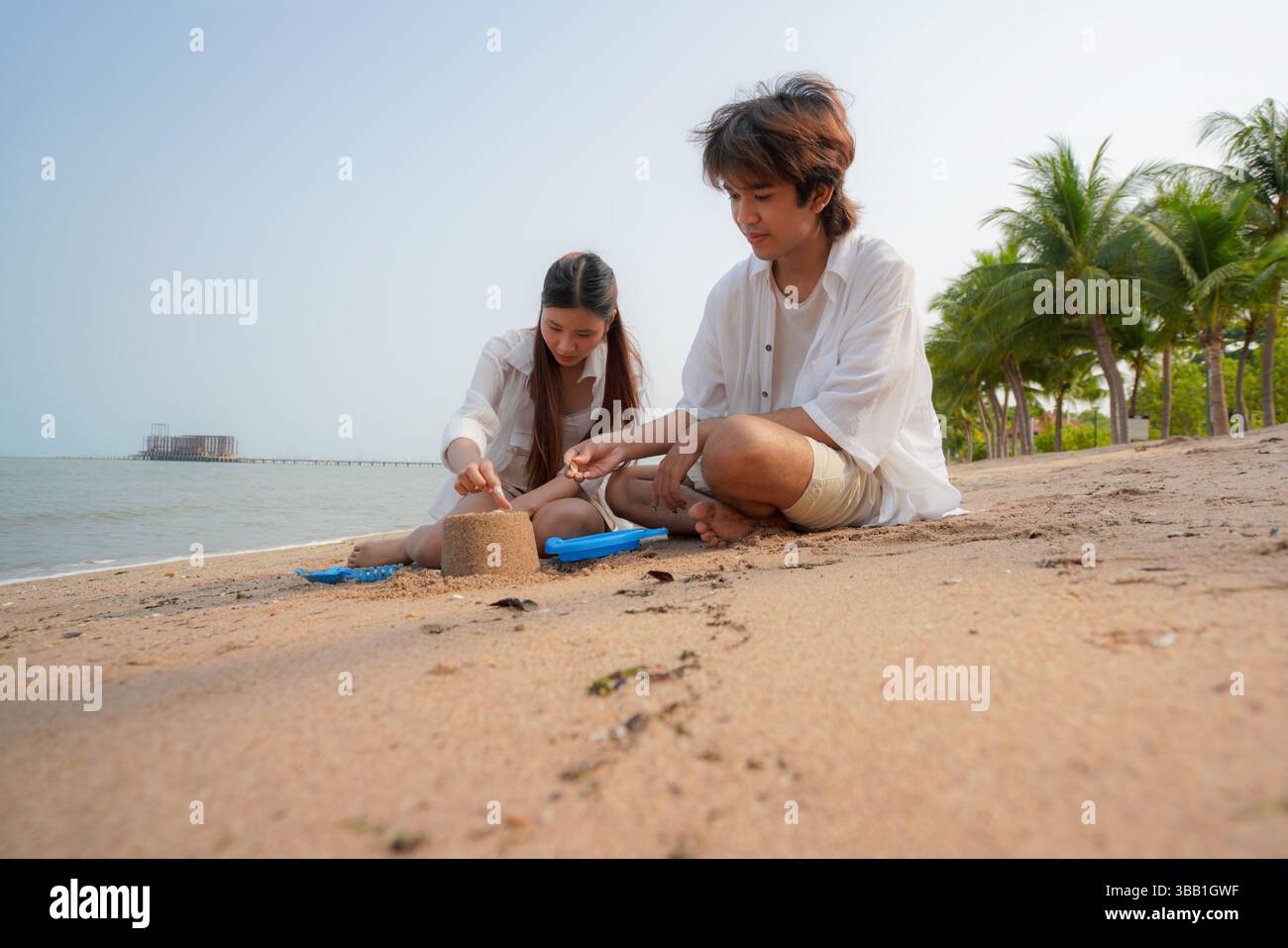 Junge asiatische Paare genießen spielerische Zeit am Strand, während sie zusammen eine Sandburg bauen, umgeben von tropischer Landschaft und ruhigem Meer, die einen SENS weckt Stockfoto