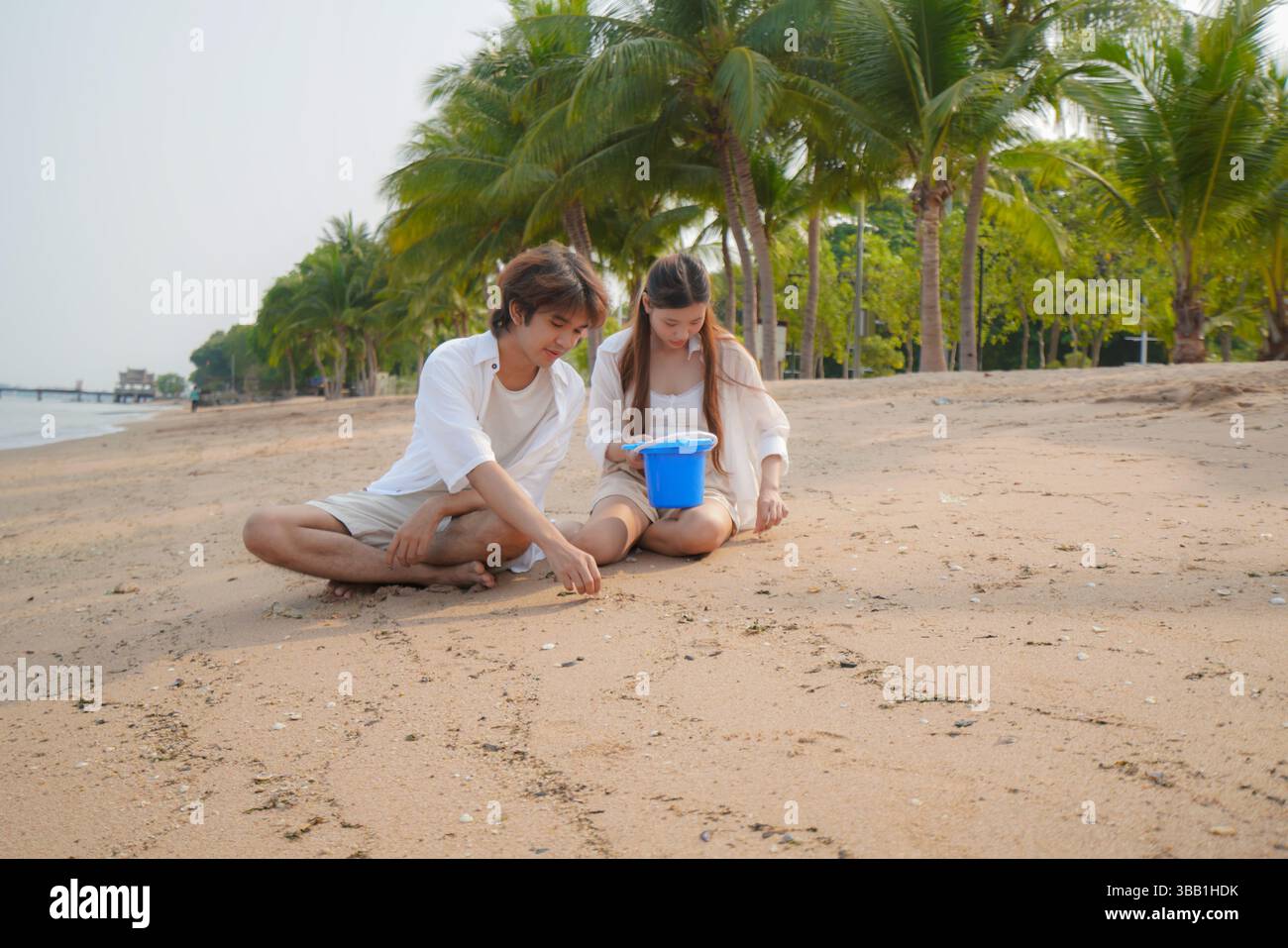 Junges asiatisches Paar, das einen ruhigen Moment am Strand genießt, während es auf dem Sand sitzt und mit einem blauen Eimer spielt, ein Gefühl der Ruhe, Nähe einfängt Stockfoto