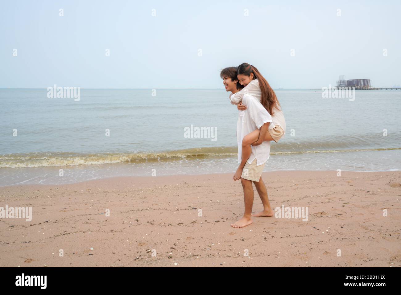 Junges asiatisches Paar verbringt schöne Zeit an einem tropischen Strand, mit dem Mann, der der Frau entlang der Küste einen Huckepack-Ritt gibt und Freude ausdrückt Stockfoto