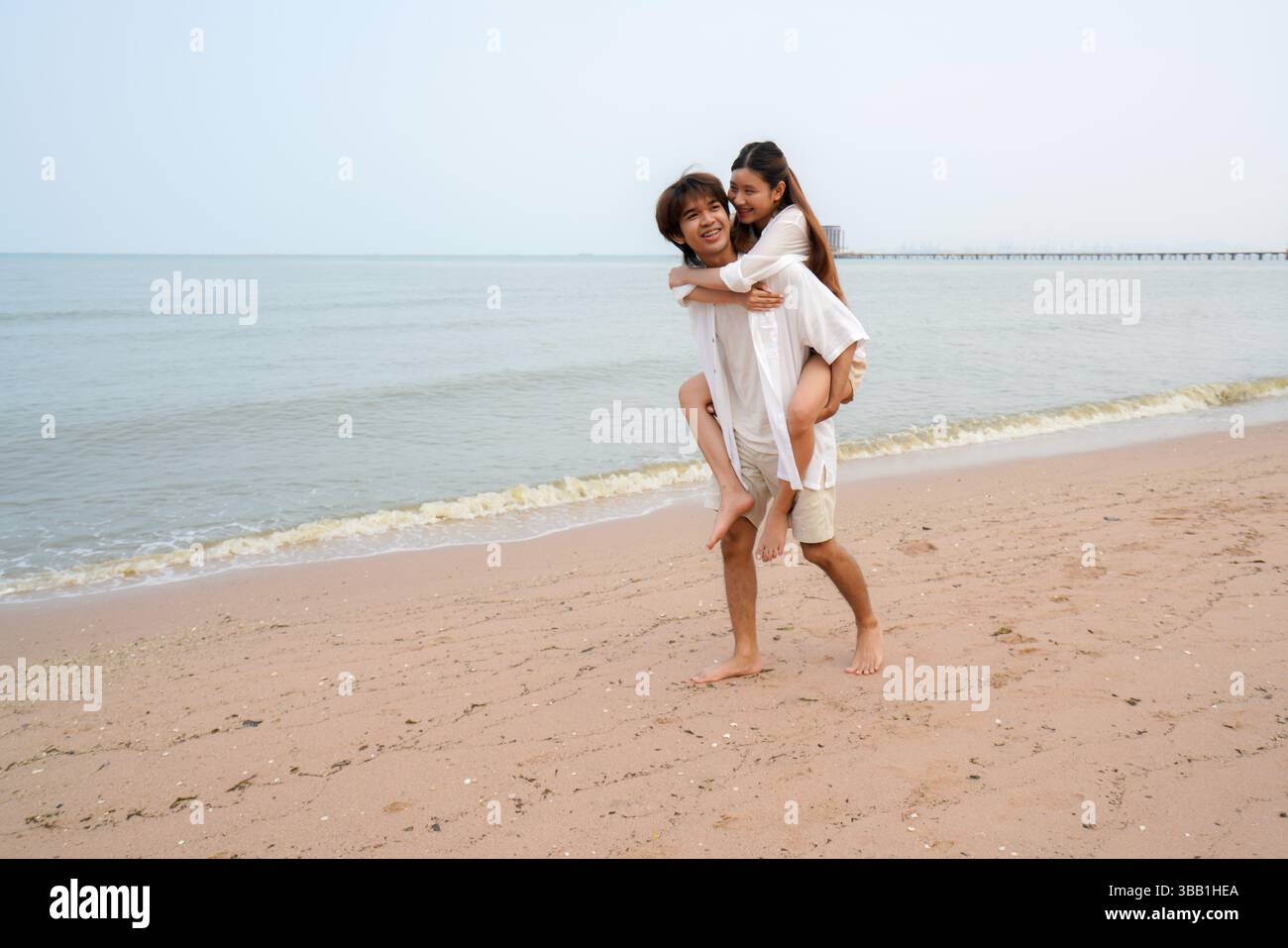 Junges asiatisches Paar verbringt schöne Zeit an einem tropischen Strand, mit dem Mann, der der Frau entlang der Küste einen Huckepack-Ritt gibt und Freude ausdrückt Stockfoto