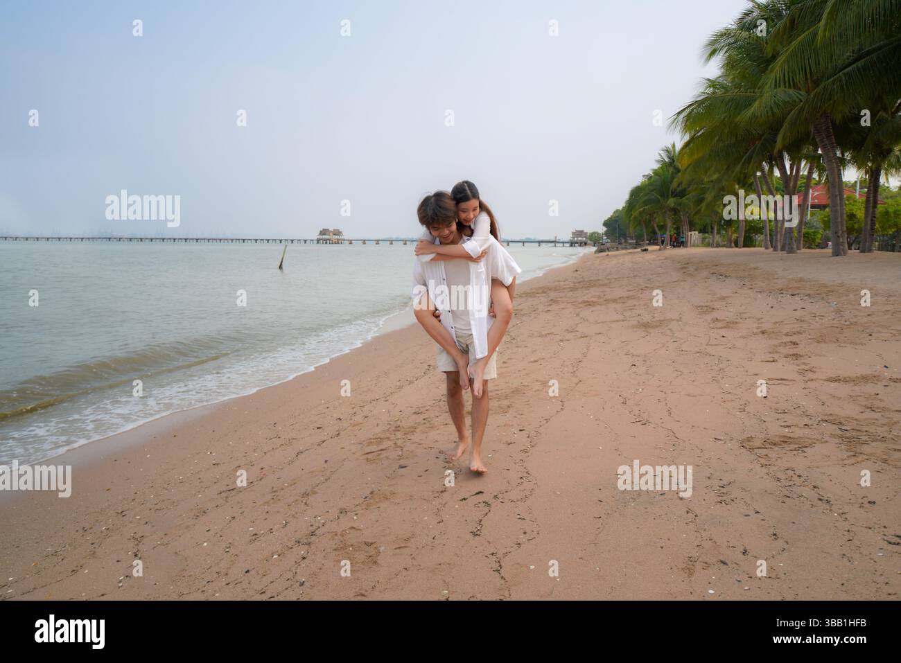Junges asiatisches Paar verbringt schöne Zeit an einem tropischen Strand, mit dem Mann, der der Frau entlang der Küste einen Huckepack-Ritt gibt und Freude ausdrückt Stockfoto