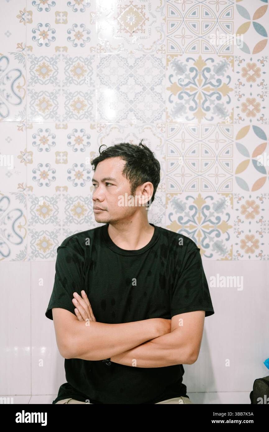 Nachdenklicher asiatischer Mann, der mit Armen an der Wand mit gemusterten Fliesen steht und auf die Seite blickt Stockfoto