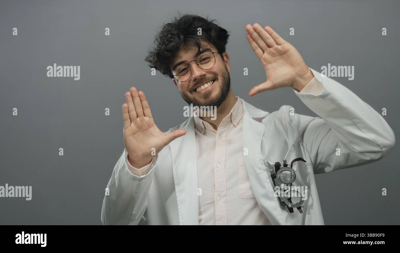 Junger hispanischer Mann mit Bart in Brille Lächeln und Gesten in einem weißen Mantel vor grauem Hintergrund, der Selbstvertrauen und Freundlichkeit zeigt. Stockfoto