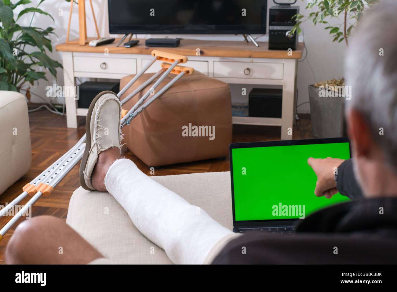 Mann mit gebrochenem Bein, der zu Hause auf dem Sofa ruht, mit Laptop mit grünem Chroma-Bildschirm Stockfoto