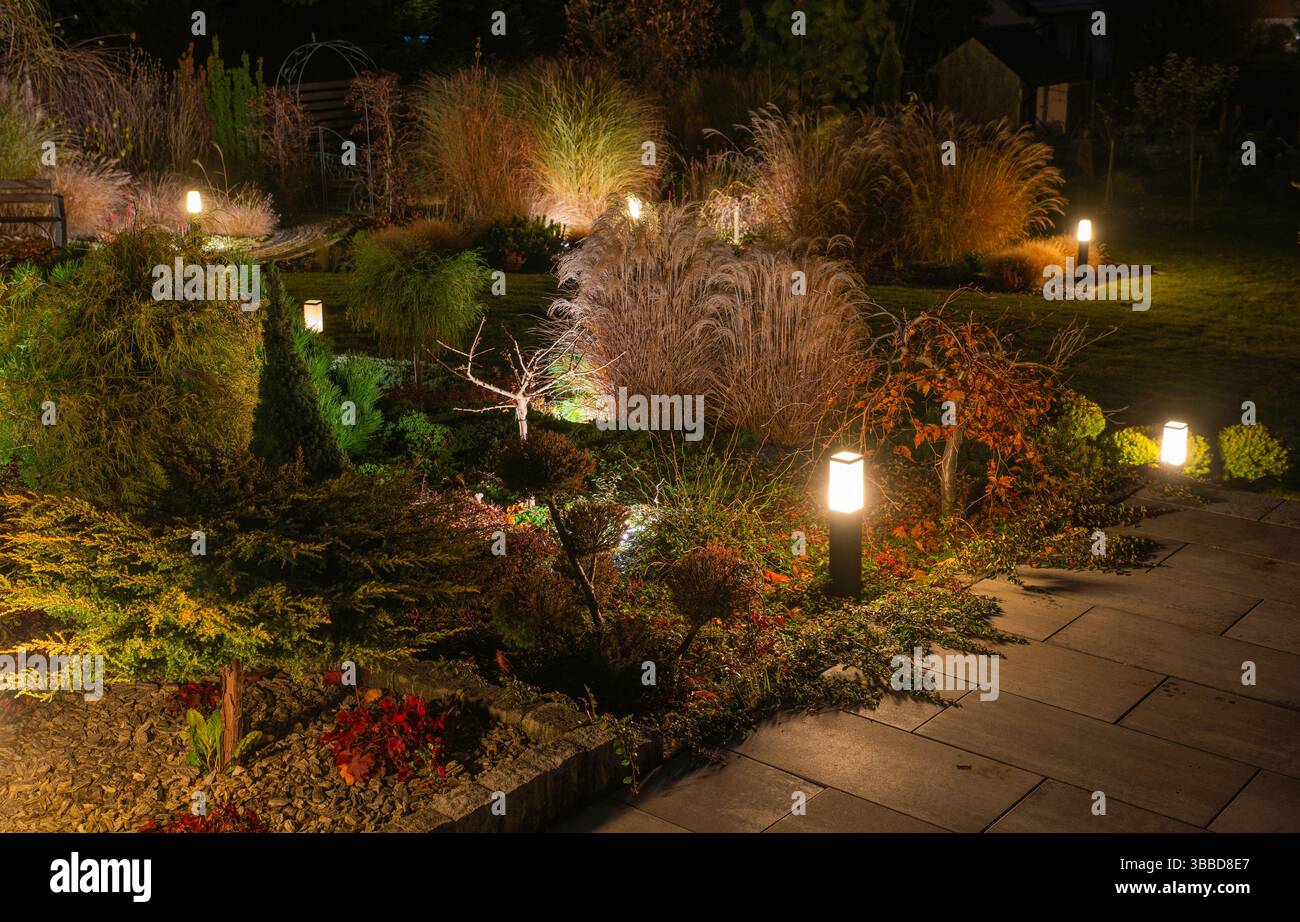 Ein wunderschön gestalteter Garten wird nachts zum Leben erweckt, mit warmen Lichtern, die verschiedene Pflanzen und Sträucher hervorheben. Dekorative Lichter leuchten sanft auf ordentlichem Glanz Stockfoto