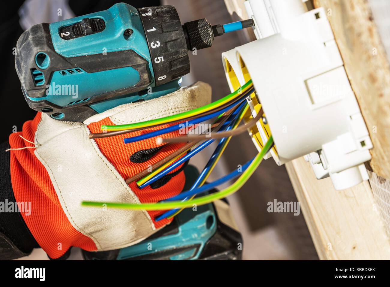 Ein gelernter Elektriker verwendet eine Bohrmaschine, um Drähte in einem an einer Holzwand montierten Anschlusskasten zu befestigen. Die elektrischen Leitungen sind in leuchtenden Farben Stockfoto