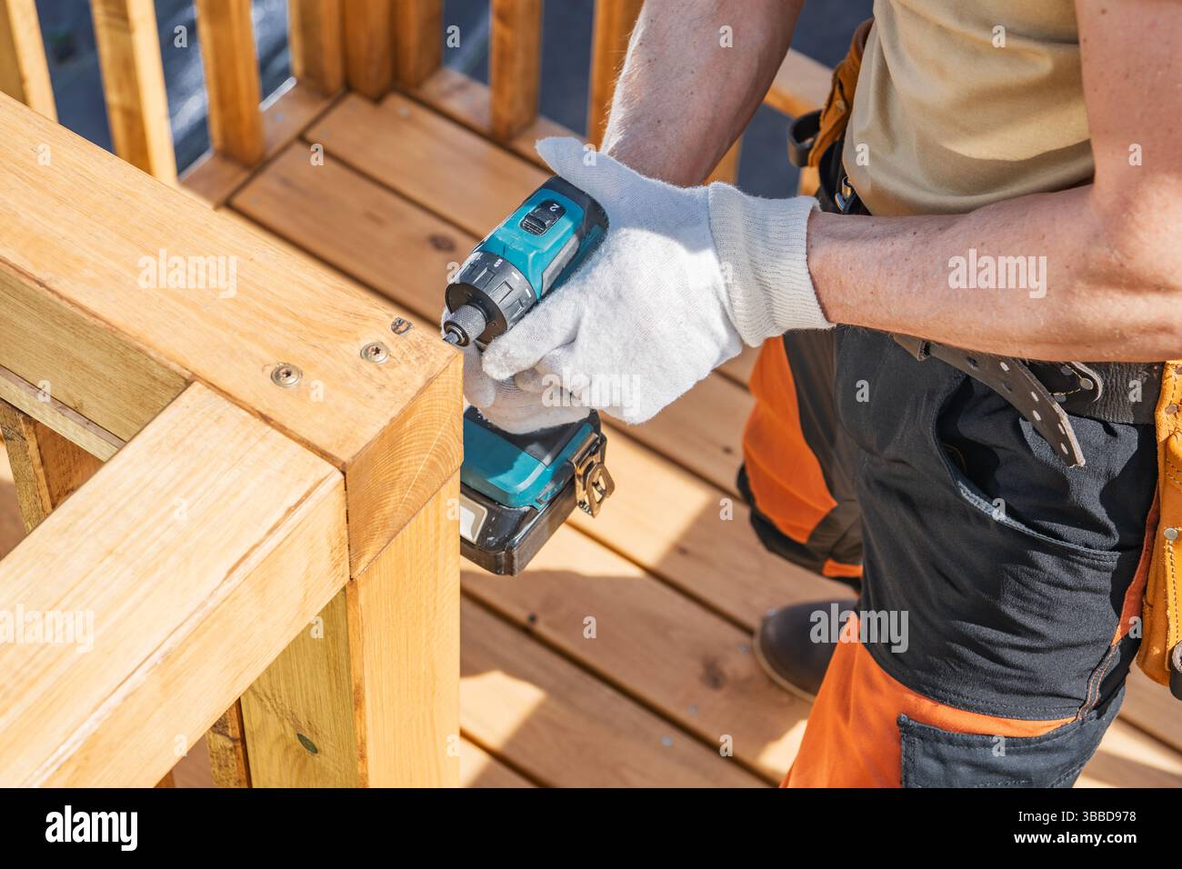 Ein Arbeiter konzentriert sich darauf, an einem hellen, sonnigen Tag ein Holzgeländer mit einer kabellosen Bohrmaschine zu sichern. Mit Handschuhen und orangefarbenen Arbeitshosen sind sie vorsichtig Stockfoto