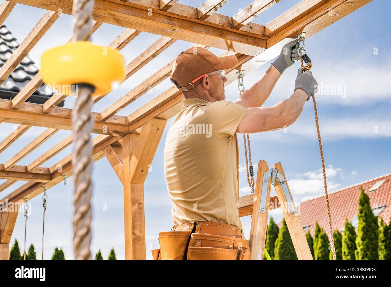 Ein Bauarbeiter konzentriert sich auf die Installation von Holzbalken an einer Außenkonstruktion und zeigt Geschick und Liebe zum Detail. Du stehst auf einer Leiter Stockfoto