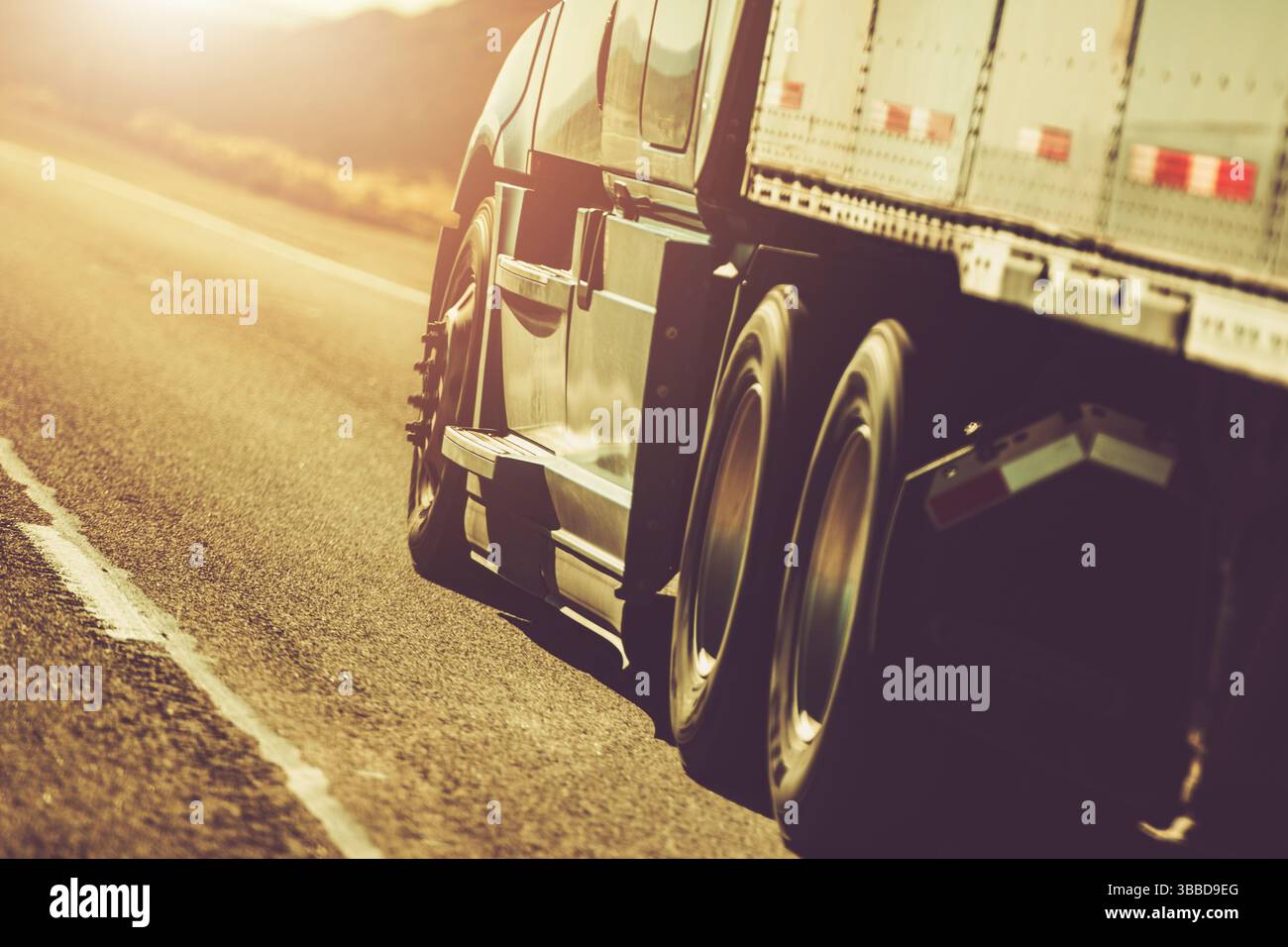 Ein großer Lkw fährt bei Sonnenuntergang über eine weitläufige Autobahn und wirft warmes goldenes Licht über die Landschaft. Der Fokus liegt auf den Hinterrädern, hoch Stockfoto