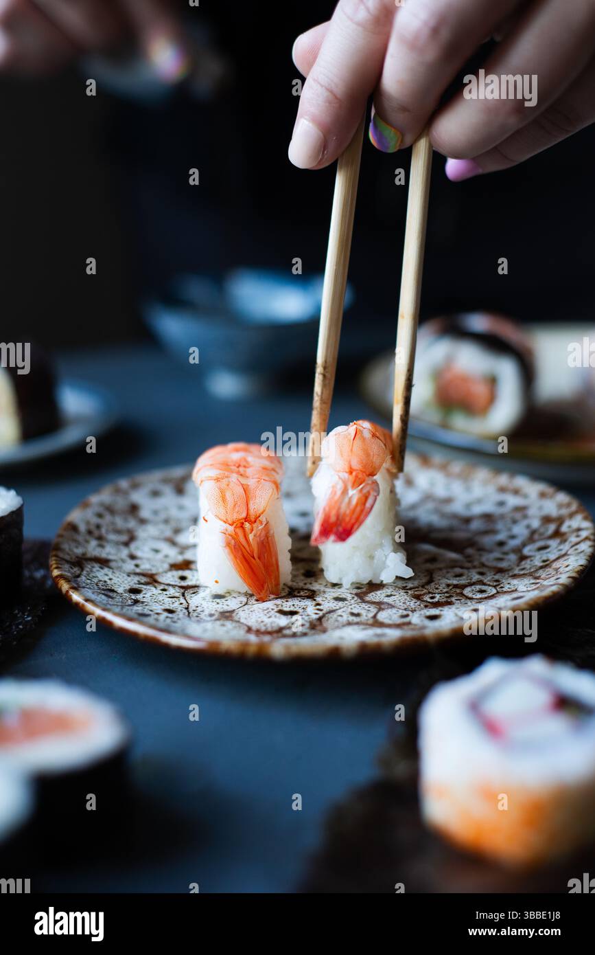 Nahaufnahme einer Person, die mit Stäbchen traditionelles japanisches ebi Nigiri Sushi mit Garnelen auf einer Keramikplatte, umgeben von verschiedenen Sushi Ro, aufnimmt Stockfoto