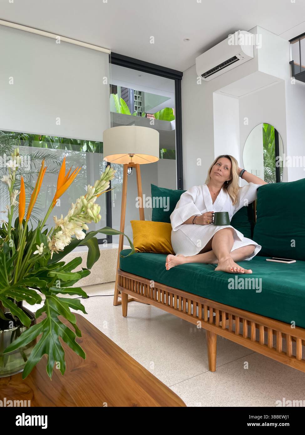 Schöne glückliche, blonde Frau mittleren Alters in einem weißen Mantel sitzt auf einem grünen Sofa mit einer Tasse Kaffee Stockfoto