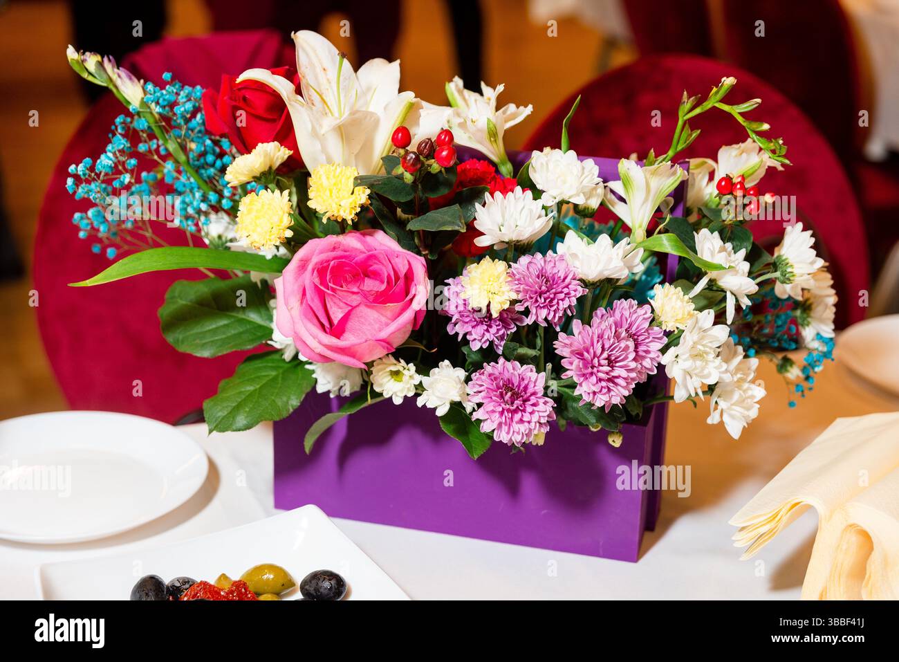 Heller Blumenstrauß mit Rosen, Lilien und Chrysanthemen in einem lila Behälter auf einem Banketttisch. Stockfoto