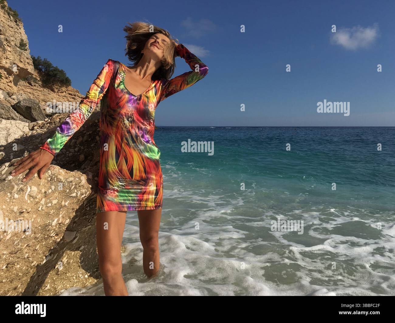 Modisches weibliches Model, das neben dem Felsen am Strand steht und mit blätternden Haaren vor dem türkisfarbenen Meerwasser auf Sardinien in Italien posiert. Schöne Frau Stockfoto