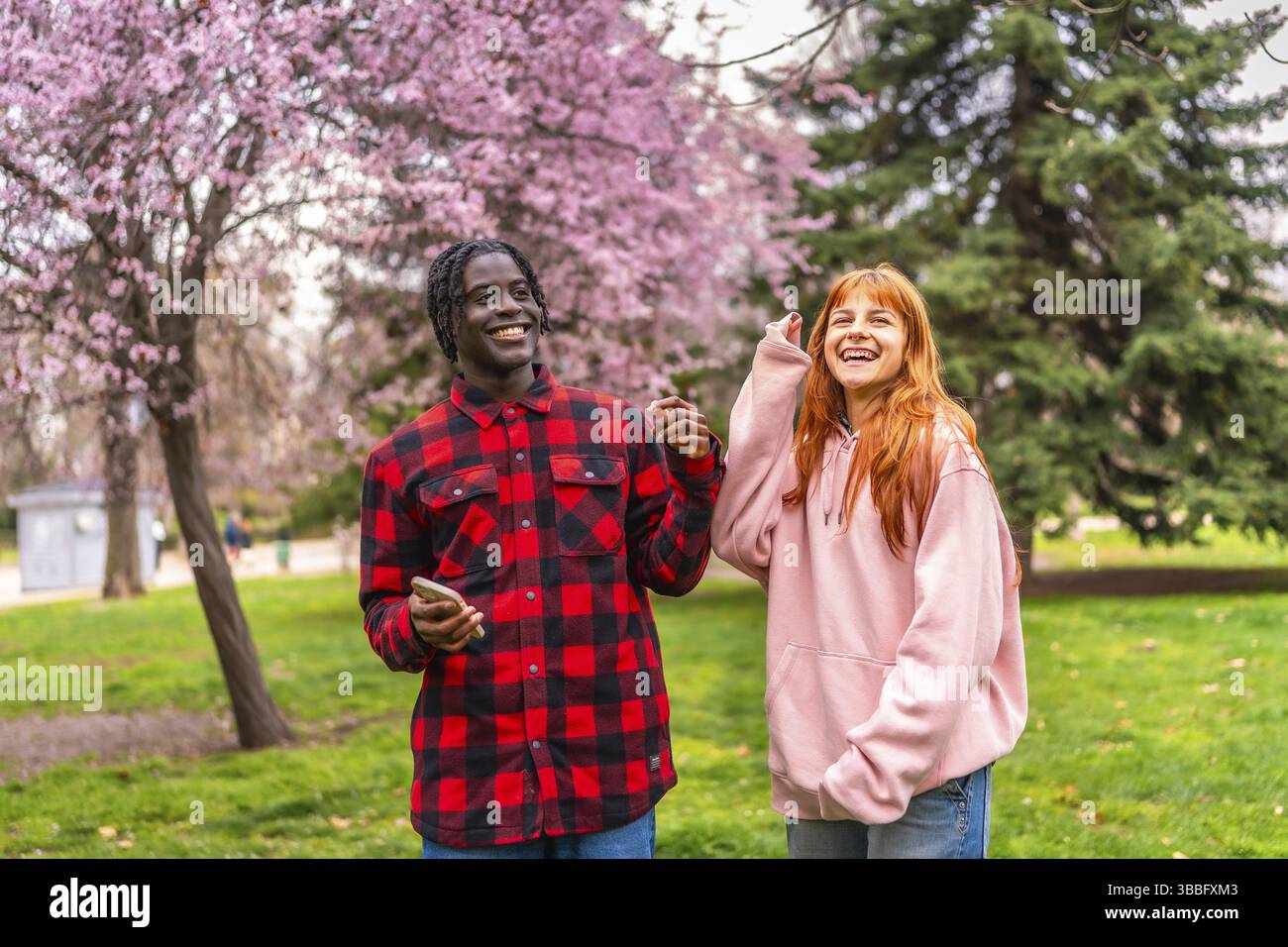 Interrassisches Paar, das einen fröhlichen Moment zusammen in einem Park voller blühender Kirschbäume genießt und die Ankunft des Frühlings feiert Stockfoto
