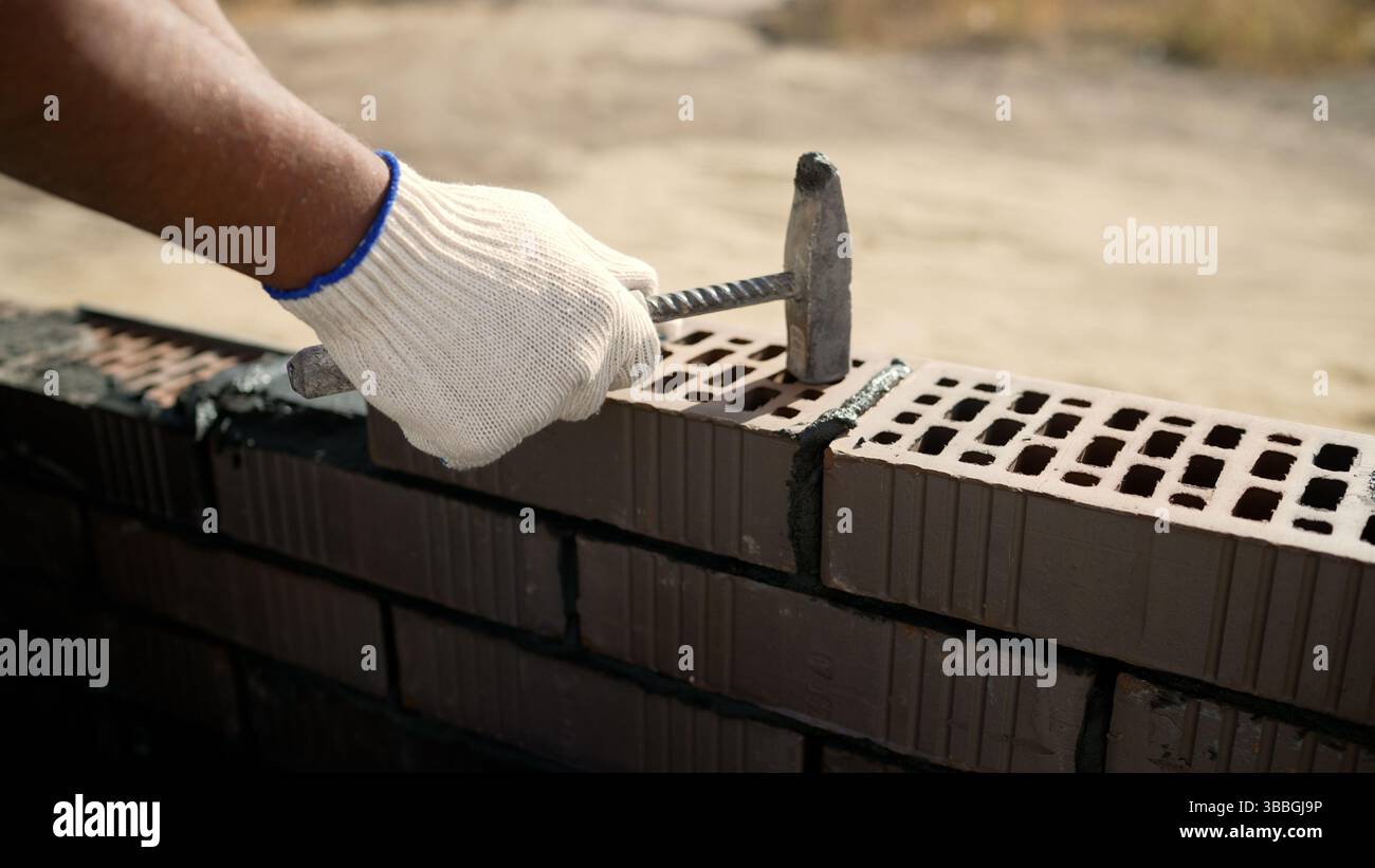 Der Bauarbeiter verwendet einen Hammer, um Ziegelsteine zu verlegen und eine Mauer zu bauen, wobei er Mörtel zwischen den Ziegeln aufträgt, um eine stabile Struktur zu erhalten Stockfoto